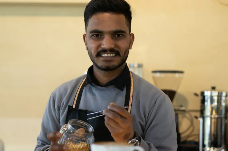 A smiling café owner in an apron behind the counter, holding a jar