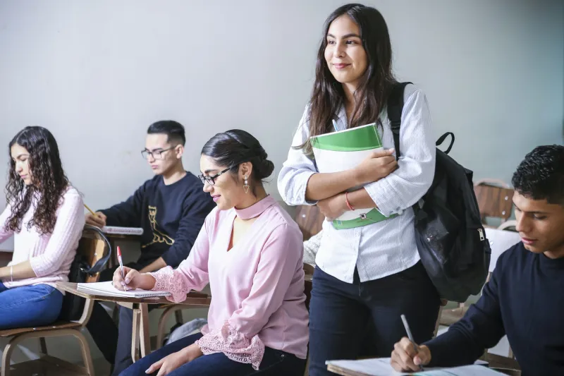 Students in a classroom; one stands smiling, holding her books