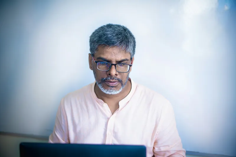 A man with grey hair and glasses working on a laptop in a white kurta