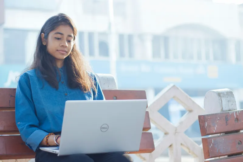 A young woman in a blue shirt working on a laptop on a wooden bench outdoors