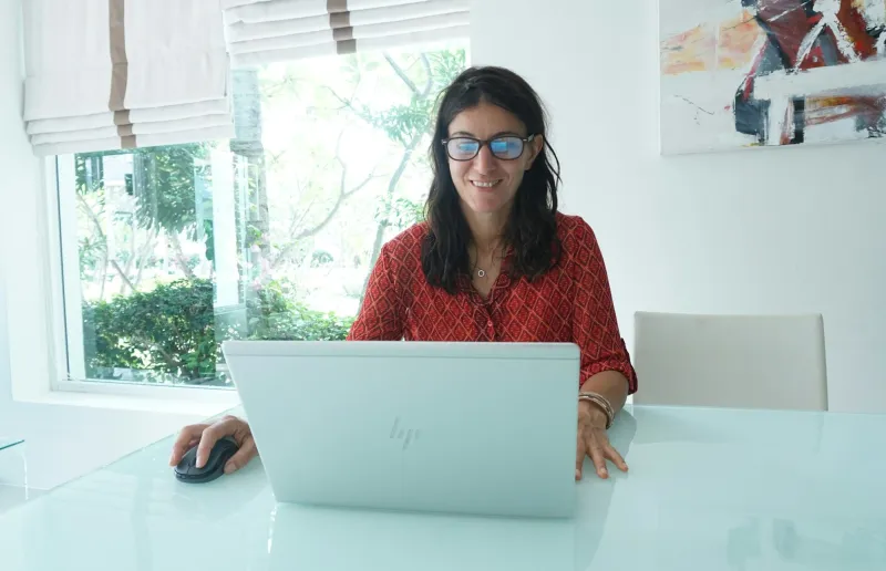 A smiling woman in a red top working on a laptop at a bright desk