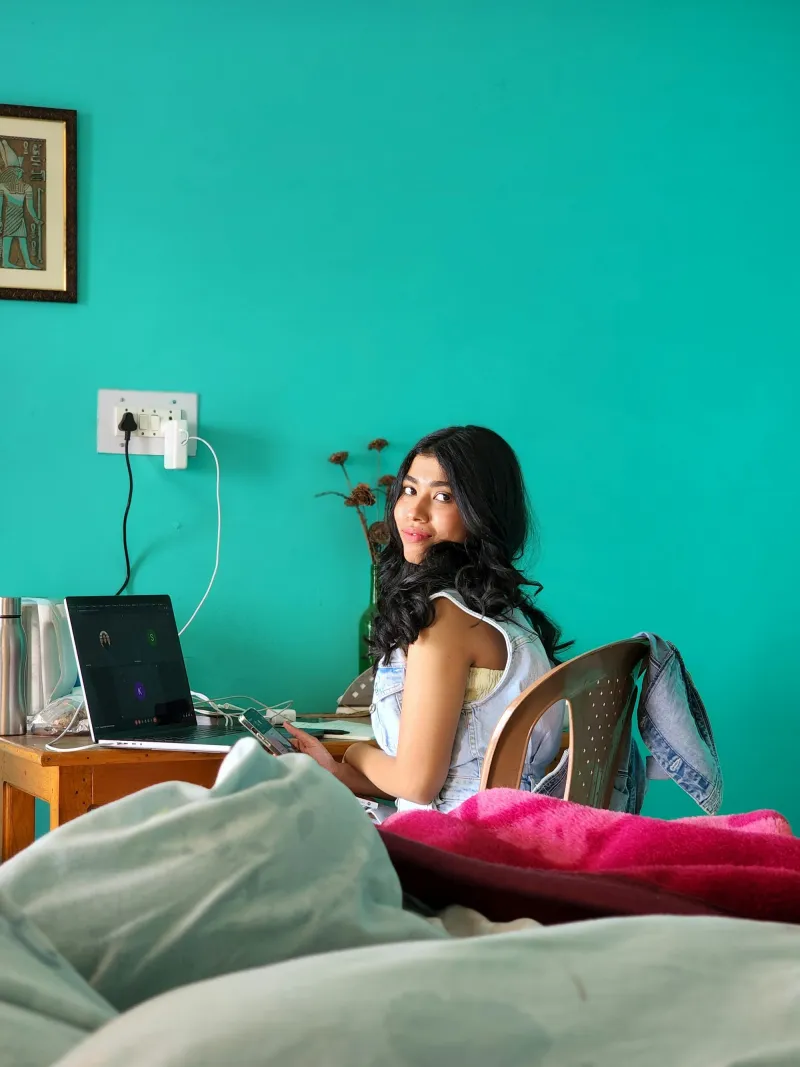 A young woman at a desk with a laptop in front of a teal wall, looking back over her shoulder