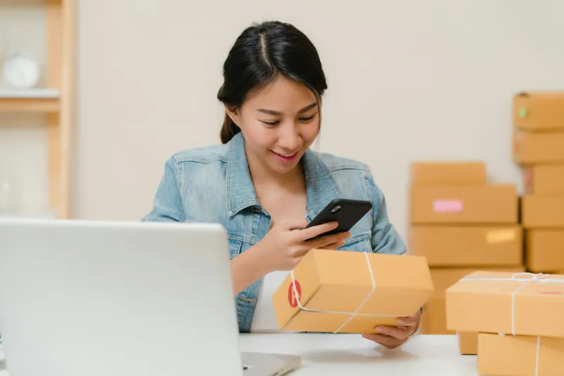 A seller checking an order on her phone beside a stack of parcels