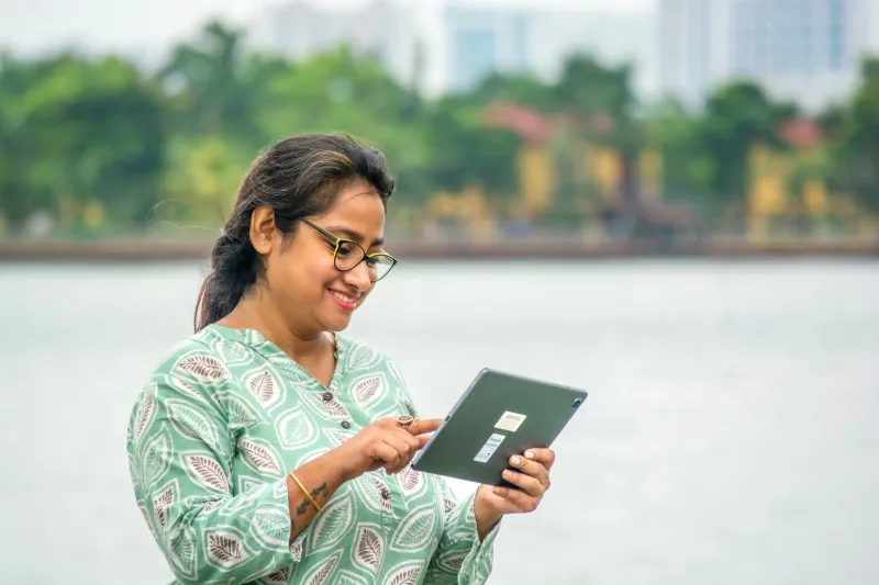 A smiling woman in a printed kurta using a tablet by a lake